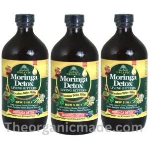 A vibrant promotional ad graphic for Essential Palace Organic Moringa Detox Living Bitters. The large 16oz dark amber glass bottle is featured prominently on the left, with a label detailing its ingredients (Lemon Grass, Fenugreek, Garlic, Juniper Berry, Fennel Extracts) and claims ("NEW 5 IN 1 Miraculous Detox Bitter"). To the right, a large white text block under a green "RELIEF FOR:" headline features a bulleted list: "Blood Sugar & Blood Pressure Levels," "Improved Digestion," "Boosts Immune System," "Enhanced Skin Health," "Energy Support," "Antioxidant, Anti-inflammatory, & Detoxify," and "Cardiovascular Health." Below the list is the website URL "www.theorganicmade.com" and a "The Organic Made" trust seal. The foreground displays fresh ingredients corresponding to the list, including lemongrass stalks, moringa leaves, and fenugreek seeds. pack of 3